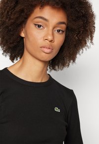 Svart ribbet langermet topp med rund hals og en liten grønn brodert krokodillelogo på brystet. Glatt tekstur.