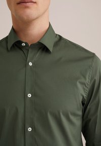 Hombre vestido con una camisa de botones color verde oliva con botones blancos, con el cuello visible, contra un fondo neutral.
