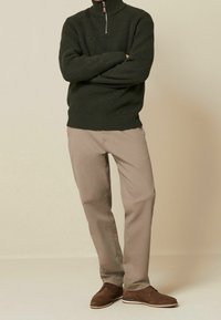 Pull en maille côtelée vert foncé avec un col zippé mi-hauteur, associé à un pantalon droit beige et des chaussures à lacets marron. Tissu texturé et coupe décontractée.