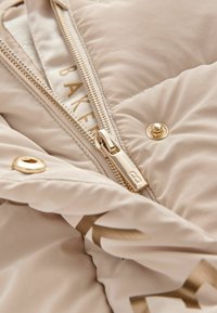 Doudoune beige clair avec quincaillerie dorée, dotée d'une fermeture éclair, d'un design matelassé et d'une doublure intérieure avec logo. Tissu texturé et finition lisse.