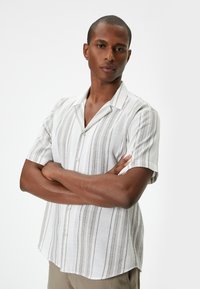 Camisa de botones de manga corta en blanco con rayas verticales grises, hecha de tejido ligero; cuenta con un cuello y un corte relajado.