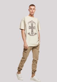 Cream oversized t-shirt with a graphic design of a cross and lion heads, paired with tan cargo pants and white sneakers.