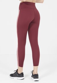 Leggings bordeaux a vita alta realizzati in un tessuto elasticizzato, con strisce laterali rosa e una texture liscia. Indossati con scarpe sportive nere.