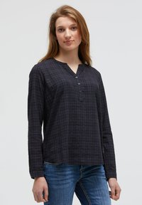 Junge Frau in einer langärmligen, marineblauen karierten Bluse mit Knöpfen und blauen Jeans, die vor einem einfachen, hellen Hintergrund steht.