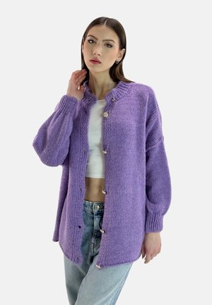 Cardigan en tricot violet avec un col montant, des manches longues et de gros boutons, associé à un haut blanc et un jean bleu clair.