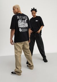 Due giovani adulti sono in piedi, uno di profilo che mostra una maglietta nera con la scritta "ROME" e una grafica del Colosseo, l'altro di fronte, con jeans neri.