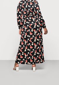 Robe maxi florale en tissu noir, avec des motifs floraux rouges et blancs, des manches longues et une ceinture à nouer à la taille. Sandales à talons blocs blanches.