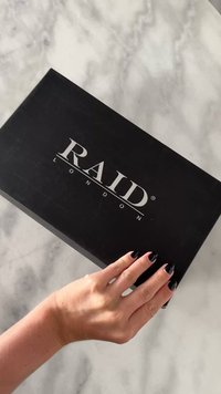 Caja negra para zapatos con "RAID LONDON" grabado en plata. Textura mate suave, sostenida con una mano, sobre una superficie de mármol.