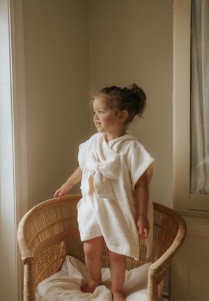 Bambino con capelli ricci in piedi a piedi nudi su una sedia di vimini imbottita, avvolto in un asciugamano bianco oversize, che guarda fuori da una finestra illuminata dolcemente.