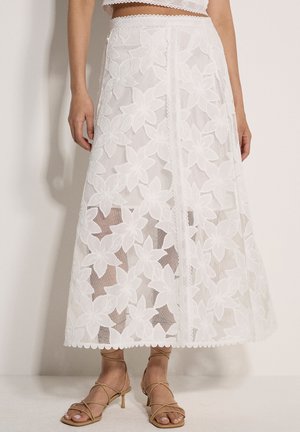 Kobieta w białej, koronkowej spódnicy midi w kwiaty i beżowych sandałach na obcasie z paskami, stojąca na tle gładkiej ściany.