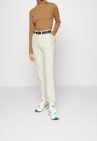 Cinturón negro, jeans rectos blancos, suéter de cuello alto a rayas marrón y naranja, y zapatillas blancas con acentos en azul y rojo. Fondo minimalista.