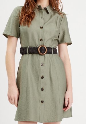 Femme portant une robe vert olive à manches courtes avec boutonnière sur le devant et une large ceinture noire tissée avec une boucle ronde en bois.