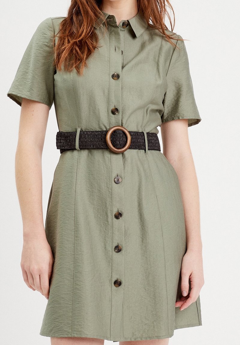 Femme portant une robe vert olive à manches courtes avec boutonnière sur le devant et une large ceinture noire tissée avec une boucle ronde en bois.