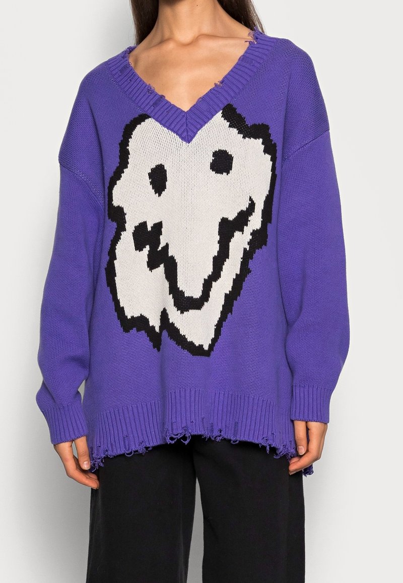Pull violet oversize col en V avec un grand motif abstrait de visage souriant blanc et noir et ourlet effiloché, porté avec un pantalon noir.