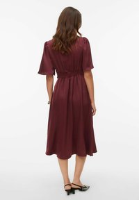 Midi haljina boje tamnocrvene s naboranim strukom, kratkim prozirnim rukavima i lepršavim rubom. Nošena s crnim cipelama sa špicastim vrhovima. Glatka satenska tekstura.