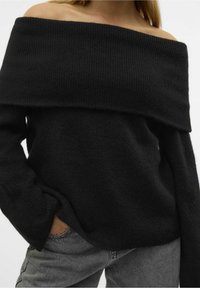 Pull noir à col bateau avec une texture en tricot côtelé épais, manches longues et coupe décontractée, associé à un pantalon gris.