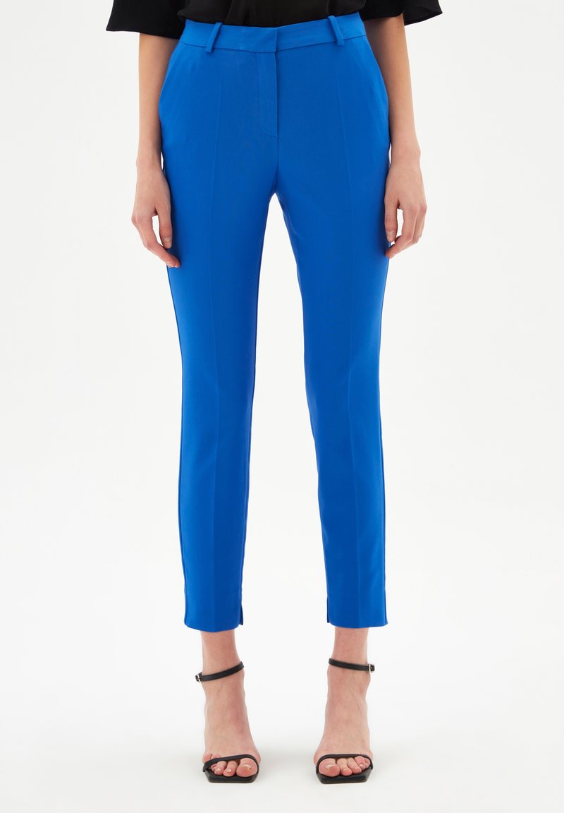 Femme portant un pantalon slim bleu vif longueur cheville avec pli devant, associé à des talons noirs à brides sur un fond clair uni.