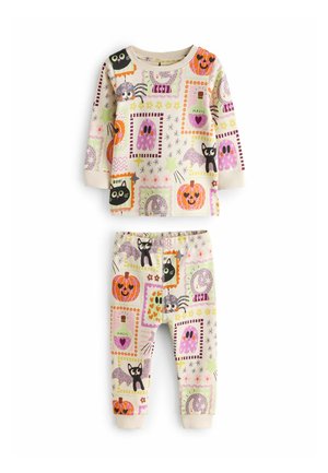 Young girl with pigtails stands indoors wearing Halloween-themed pajamas with cats, pumpkins, spiders, ghosts, and stars on a light background.