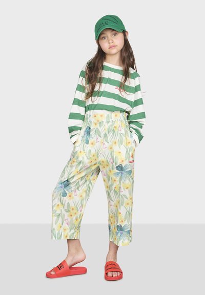 Camicia a maniche lunghe a righe verdi e bianche, pantaloni larghi con stampa floreale, sandali rosso a scivolo e un cappello verde con testo.