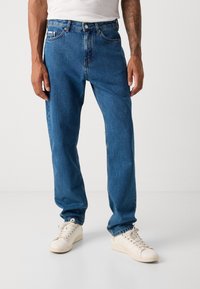 Mann som har på seg blå denimjeans, hvite joggesko og en hvit T-skjorte, står mot en enkel lys bakgrunn.
