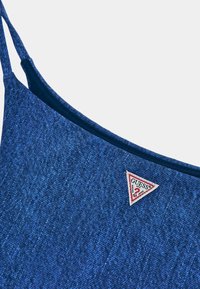 Jeansblus i blått med tunna axelband, med en triangulär röd och vit Guess-logotyp-lapp. Strukturerat tyg med en lätt lyster.