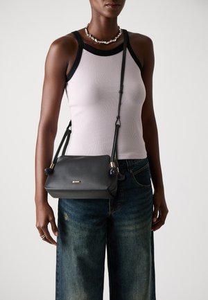 Sac en bandoulière en cuir noir avec des garnitures en or, bords arrondis et un fermoir simple. Porté sur un débardeur rose clair et un jean déchiré.