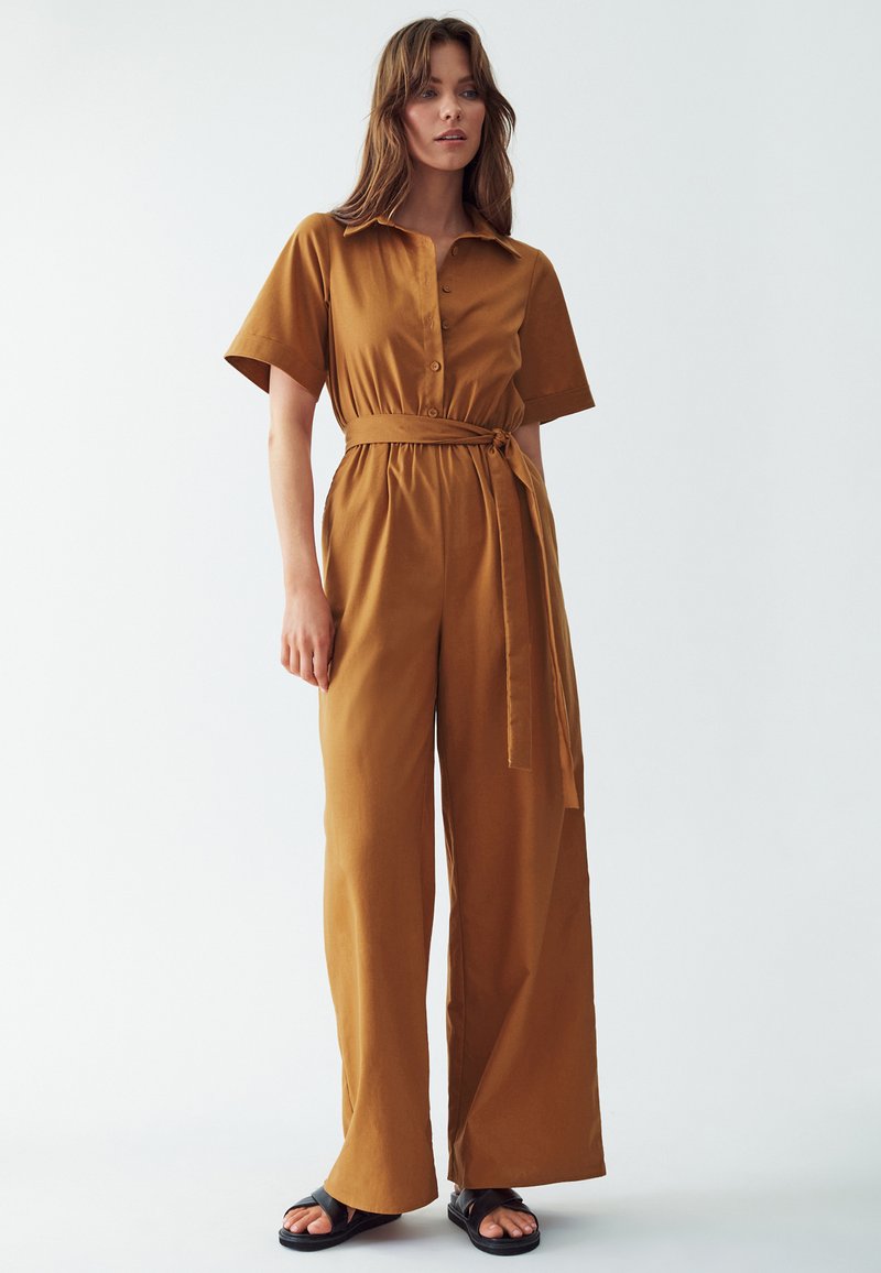Hnědý overal s krátkým rukávem a zapínáním na knoflíky, s přiléhavým pasem a páskem na zavázání, a širokými nohavicemi. Stylizováno s černými klouzavými sandály.