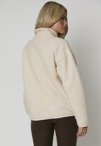 Kvinna med blont hår som bär en krämfärgad, strukturerad fleecejacka sedd bakifrån, tillsammans med mörkbruna byxor mot en enfärgad bakgrund.