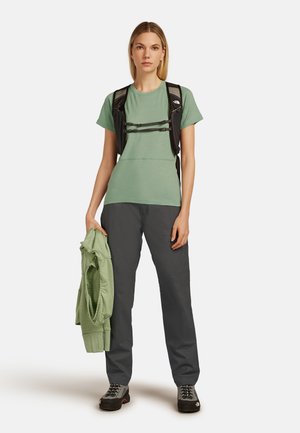 Helles grünes Kurzarmhemd, graue Hose und dunkler Rucksack. Trägt schwarze Schuhe. Hält einen passenden grünen Kapuzenpullover.