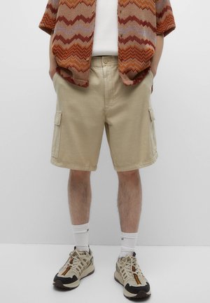 Shorts cargo beiges avec deux poches latérales, associés à une chemise tricotée à manches courtes à motifs et des baskets beiges. Chaussettes blanches unies visibles.