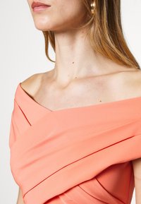 Off-the-shoulder coral dress featuring draped fabric, soft texture, and diagonal pleats. Accented with a slight sheen and minimalistic design.