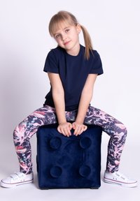 Dziecko w granatowej koszulce siedzi na granatowym aksamitnym sześcianie. Różowe legginsy w kwiaty i białe trampki z podwójnymi paskami. Proste tło.