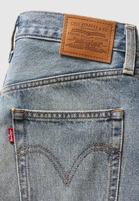 Lichtblauwe denim jeans met een bruine leren patch met verhoogde branding, contrasterende oranje stiksels en een rode Levi's tab op de achterzak.