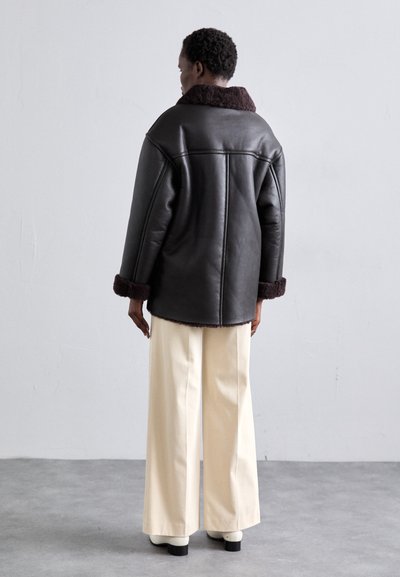 Prevelika crna kožna jakna s smeđim ovratnikom i manšetama od ovčjeg runa, uparena s kremastim hlačama širokih nogavica i glomaznim bijelim potpeticama.