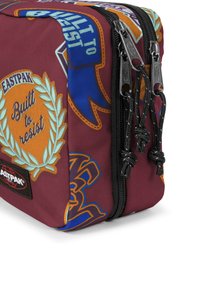 Burgundská taška Eastpak s barevnými nášivkami, která má design listu a dva černé zipové táhla s pletenými šňůrkami.