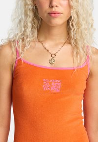 Πορτοκαλί τοπ χωρίς μανίκια από μαλακό ύφασμα με ροζ τιράντες. Διαθέτει το λογότυπο "BILLABONG" και ροζ κεντήματα με λουλούδια στην περιοχή του στήθους.