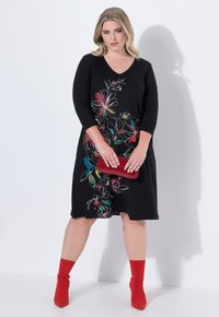 Robe noire avec broderies florales colorées, manches trois-quarts, décolleté en V, accompagnée d'une pochette rouge et de chaussettes de cheville assorties.