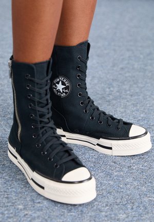 Czarne wysokie trampki Converse z białymi gumowymi podeszwami i noskami, noszone na stopach na szarej teksturowanej powierzchni.