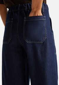 Tmavě modré džínové kalhoty s volným střihem, elastickým pasem a viditelnými zadními kapsami. Kontrastní žluté švy zvýrazňují design.