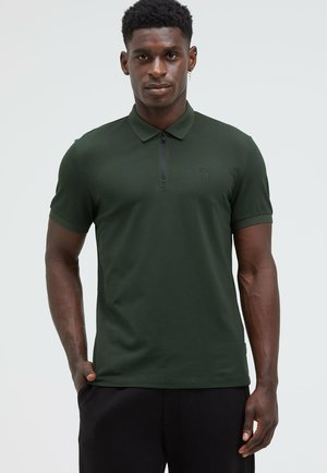 Dark green polo shirt with a zip collar, short sleeves, and a subtle logo on the chest. Smooth, lightweight fabric with a fitted design.