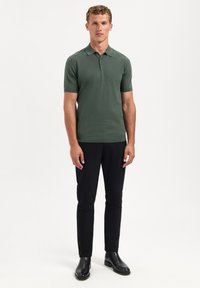 Groene polo met korte mouwen en een kraag, met een sluiting van drie knopen en een gladde textuur; gecombineerd met zwarte slim-fit broeken en zwarte laarzen.