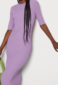 Robe violette moulante à côtes, manches courtes et encolure ronde. Texture lisse. Portée avec un sac à main vert pour le contraste.