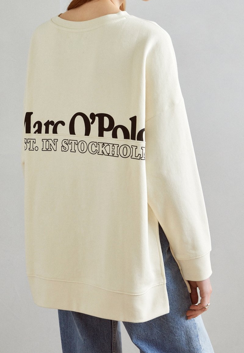 Personne portant un sweat-shirt crème avec le texte « Marc O’Polo EST. À STOCKHOLM » dans le dos, associé à un jean bleu.