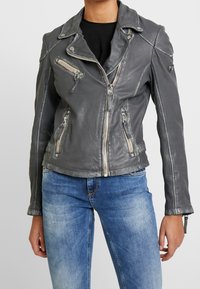 Kvinde iført en tætsiddende grå læder bikerjakke med lynlåslommer og blå slidte jeans foran en ensfarvet baggrund.
