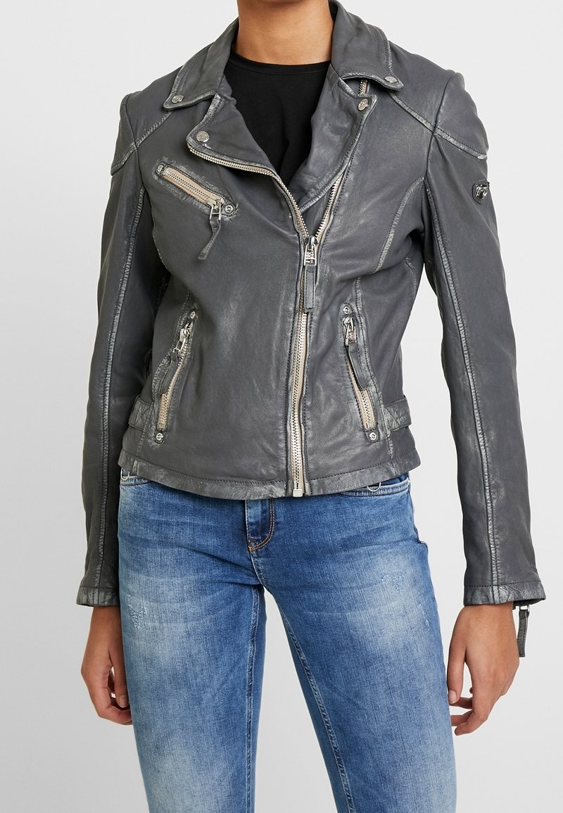 Kvinde iført en tætsiddende grå læder bikerjakke med lynlåslommer og blå slidte jeans foran en ensfarvet baggrund.