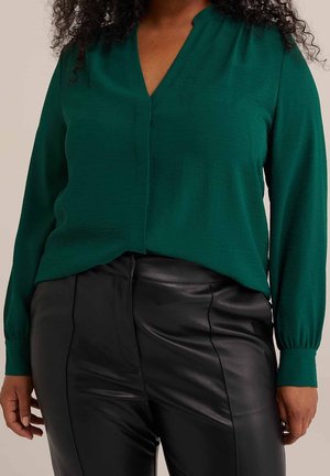 Mujer con una blusa verde de manga larga metida en pantalones de cuero negros de talle alto, de pie frente a un fondo neutro.