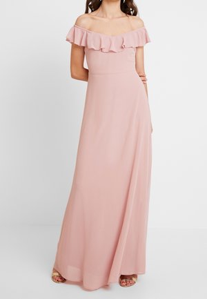 Vrouw draagt een lang, roze jurk met blote schouders, een gerimpelde halslijn en open sandalen en staat met één arm achter haar rug.