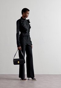 Čierna blúzka s volánmi a gombíkmi, v kombinácii s čiernymi šitými nohavicami. Doplnková malá čierna kabelka s okrúhlym zlatým uzáverom.