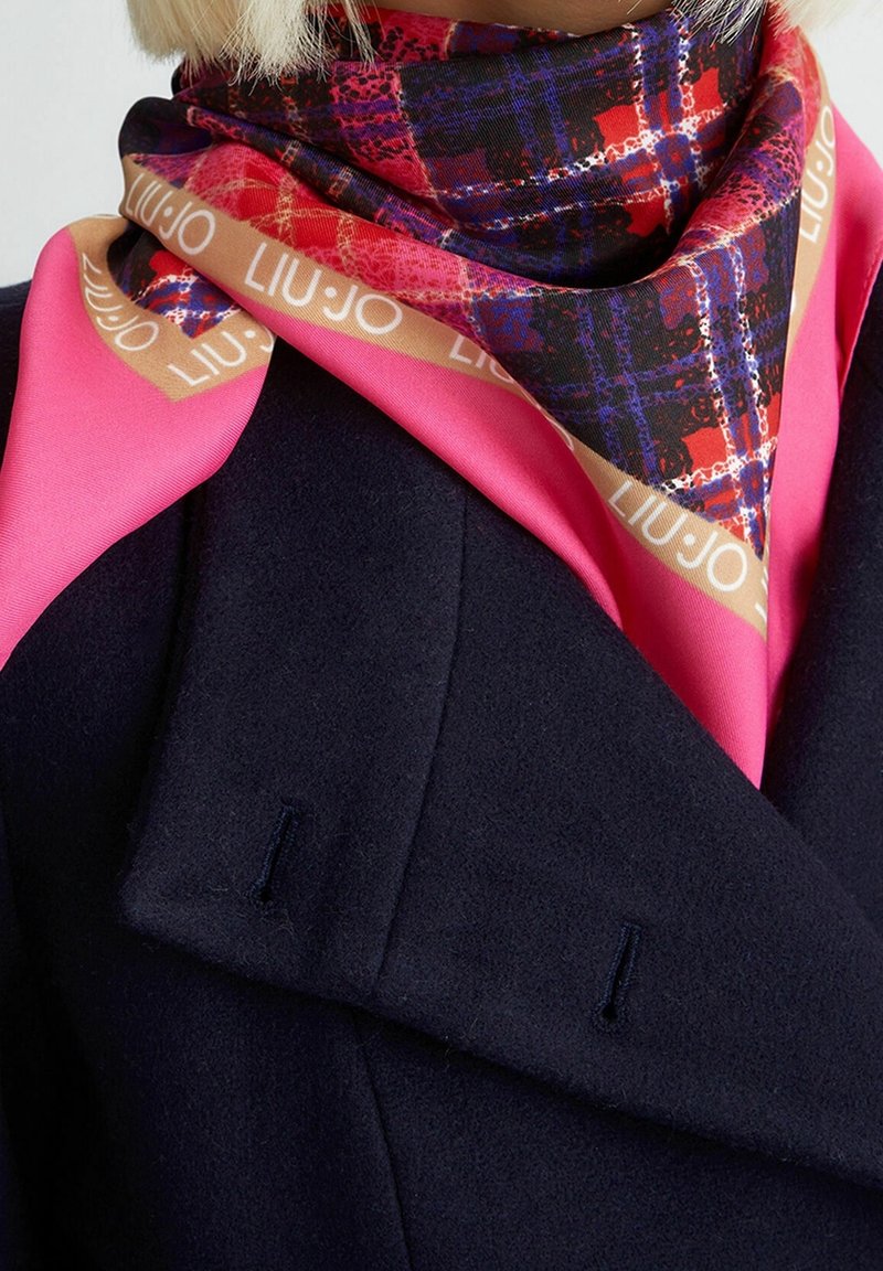 Femme portant une écharpe à carreaux multicolores avec un texte beige « LIU·JO » sur un top rose vif et un manteau bleu marine foncé.