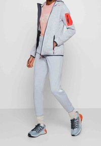 Grijze fleecejack met een rode ritszak, zwarte afwerking, roze t-shirt, lichtgrijze broek en blauwe sneakers met oranje accenten.